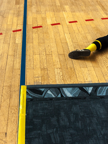 Der Unterschenkel eines Spielers beim Blindensport Torball liegt auf dem Hallenboden vor einem Markierungs-Teppich