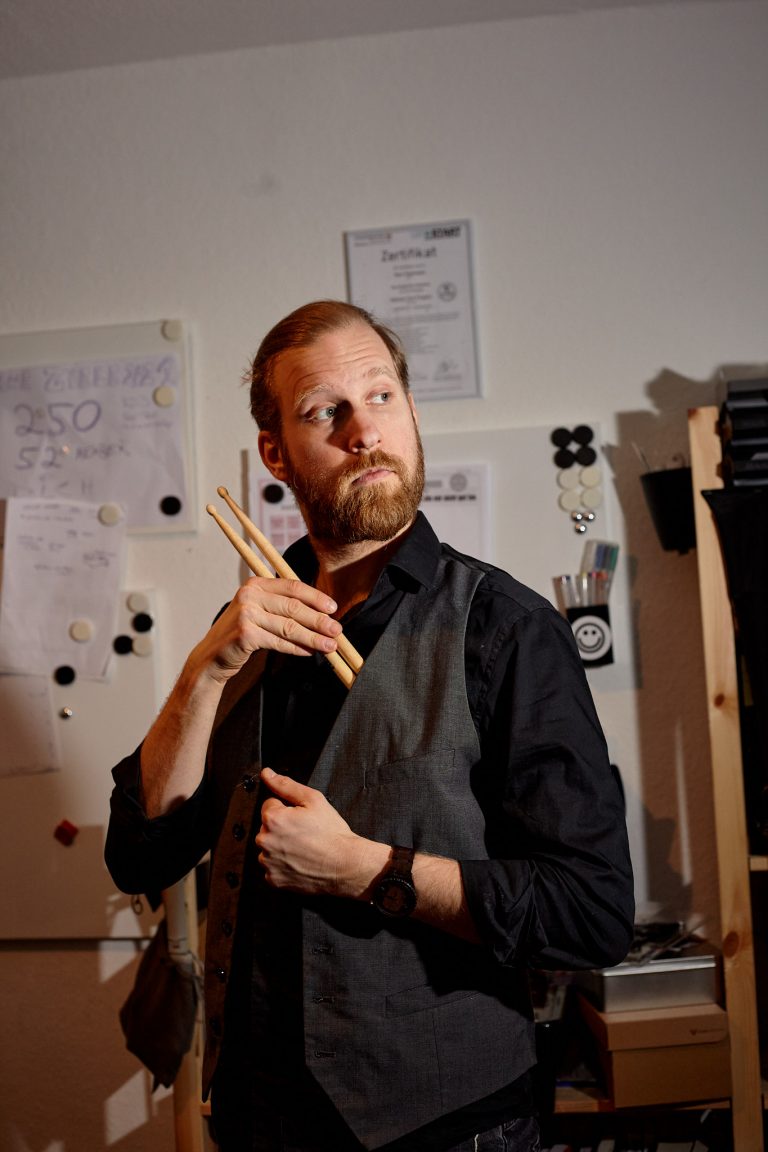 Portrait von Marc Pawlowski
| Musiker in seinem Arbeitszimmer, Schlagzeugsticks haltend.