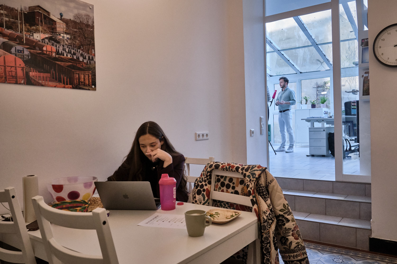 Eine Mitarbeiterin des Wahlkreisteams von Felix Banaszak, Bundestagsabgeordneter für Bündnis 90/Die Grünen, arbeitet an ihrem Laptop an einem Tisch im Wahlkreisbüro. Im Hintergrund gibt der Abgeordnete einem TV-Sender ein Interview.A staff member of the constituency team of Felix Banaszak, Member of the Bundestag (Germany’s federal parliament) for The Greens, works on her laptop at a table in the constituency office. In the background, the parliamentarian is giving an interview for a TV broadcaster.