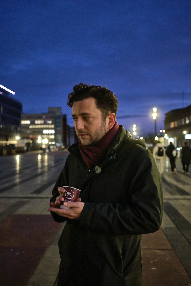 Felix Banaszak, Bundestagsabgeordneter von Bündnis 90/Die Grünen, steht frühmorgens am Duisburger Hauptbahnhof und wärmt sich kurz bei einem Kaffee auf. Dort unterstützte er den Wahlkampf seiner Partei, indem er Wahlkampfflyer verteilte.

Felix Banaszak, a Member of the Bundestag (Germany’s federal parliament) for Bündnis 90/Die Grünen (The Greens), stands at Duisburg Central Station early in the morning, warming up briefly with a coffee. There, he assisted in his party’s election campaign by handing out campaign flyers.