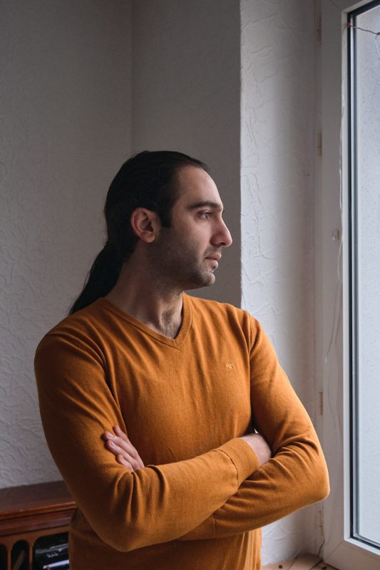 Portrait von Roozbeh in seiner Wohnung in Dortmund. Roozbeh gehört zur iranischen Diaspora in Deutschland. Aufgenommen am 03.12.2022 in Bochum.

Roozbeh kommt aus 
Sanandaj in den kurdischen 
Gebieten im Westen Irans
Er ist Musiker und Produzent, spielt Santour (ein zitherähnliches iranisches Instrument) und Geige und produziert überwiegend Musik für
 die internationale 
kurdische Community. 
In der Vergangenheit wurde ihm wiederholt mit Entführung gedroht, sollte er seine musikalische Tätigkeit für verschiedene kurdische Parteien nicht einstellen. Er floh 2015 nach Deutschland. Inzwischen ist sein Status als politischer Flüchtling in Deutschland
 offiziell anerkannt. 
Während der Übergriffe der Revolutionsgarden auf die Bevölkerung in seiner Heimatstadt Sanandaj Anfang Oktober 2022, sah er in den sozialen Medien ein Video auf dem die Schüsse der Sicherheitskräfte auf die Zivilbevölkerung zu sehen und zu hören waren.  Das Video war von einem Aussichtspunkt aufgenommen, von dem Roozbeh selbst oft auf seine Heimatstadt geschaut hatte. Sie nun so zu sehen, war ein sehr schmerzhafter, verzweifelter
 Moment für ihn. 

Zitat:"Große Teile der Bevölkerung haben die Hoffnung auf Besserung verloren, sie haben nichts mehr zu verlieren. Entweder leben wir in Würde, oder wir leben gar nicht. Es  gibt da diese Mentalität in einer Revolution zu sein und es gibt keine Anzeichen, dass das nachlassen wird. Die Protestierenden lernen dazu, kommunizieren und entwickeln Strategien. Niemand im Iran ist noch dieselbe Person wie vor drei Monaten. Die Menschen sind sehr entschloßen und wollen nicht zur Normalität zurückkehren."